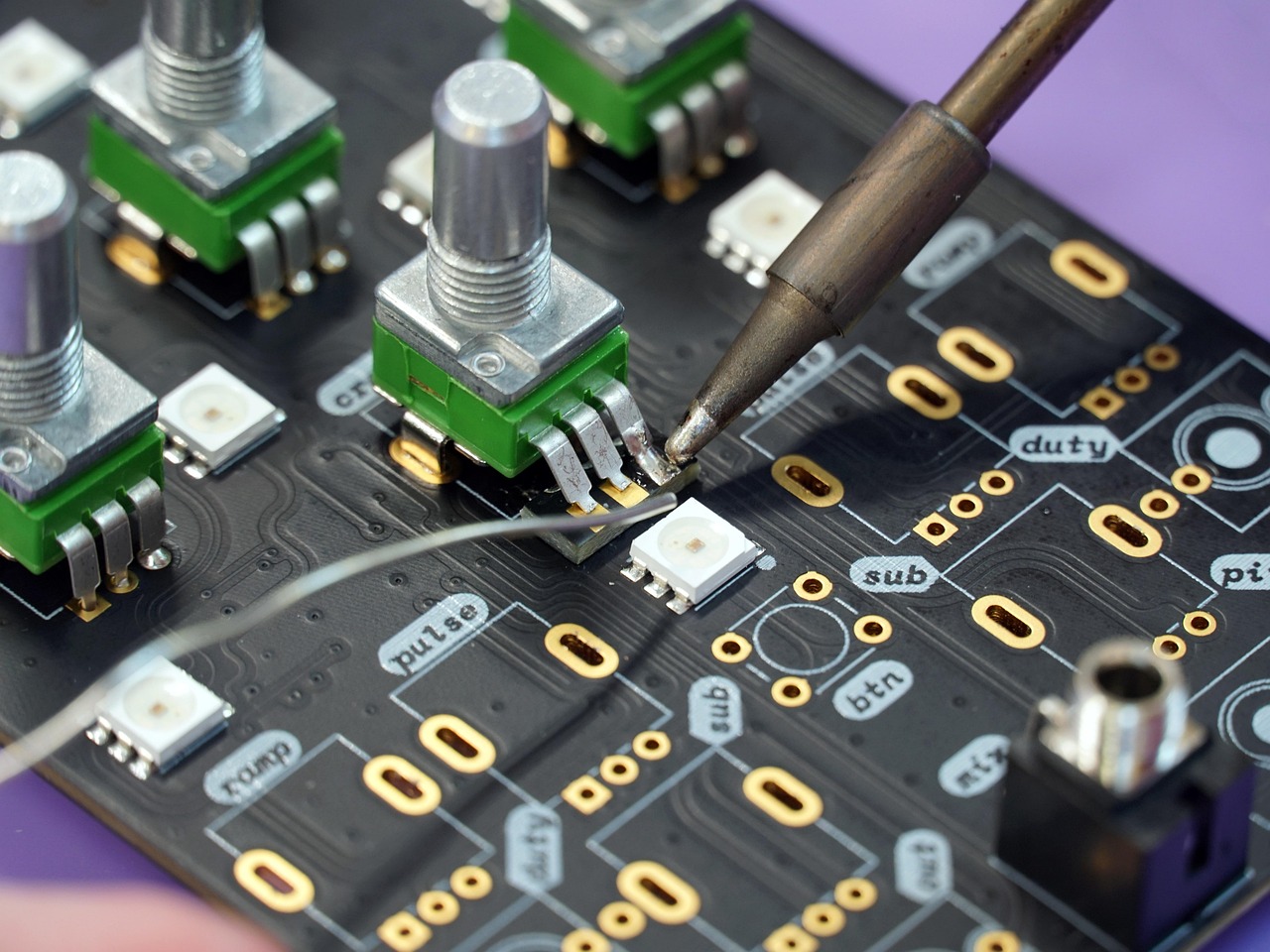 Close-up of a black circuit board with green potentiometer bases being soldered